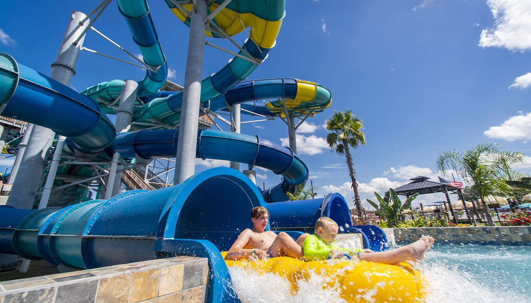 BLOG Two children on a yellow inflatable raft exit a blue water slide into a splash pool at a water park under a clear blue sky.